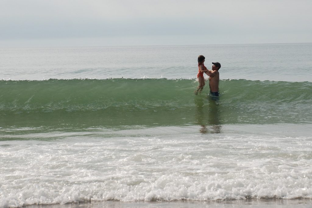 Image of a wave breaking in the ocean with two figures to the right, a father and a daughter. The father, in a baseball cap, is lifting the daughter over the wave as it breaks. The foam of earlier waves are in the foreground. It is a relatively overcast day.