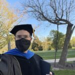 Chris Long in graduation regalia and mask in front of a leafless Resilient Tree.