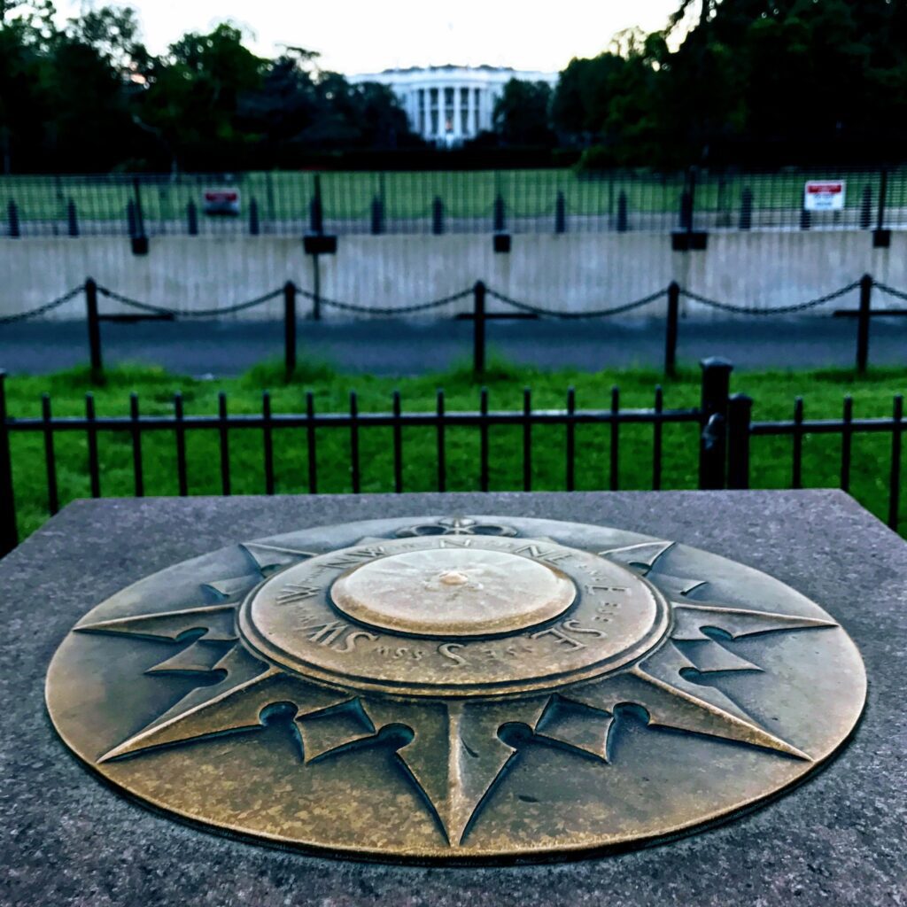 Compass with directions in the foreground with a distant and unfocused image of the White House in the distance.