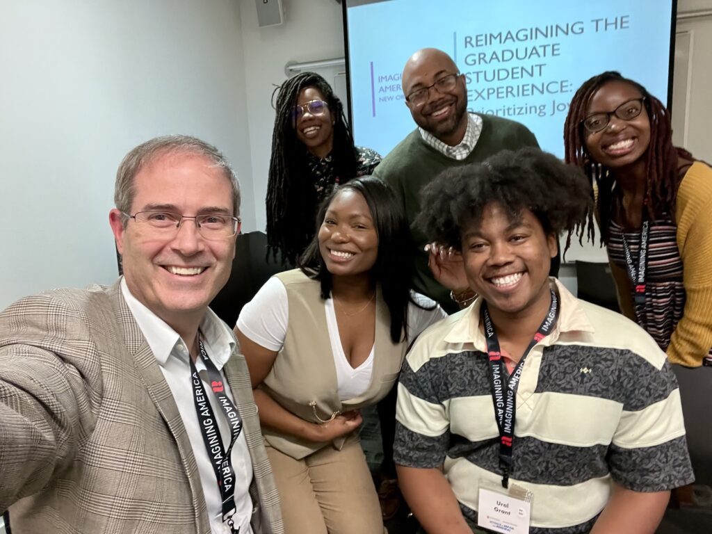 Selfie with Chris Long on the left Toni Gordon and Ural Grant in the front row and behind them from left to right, Sharieka Botex, Marquis Taylor, and Jessica Reed. We are standing in front of a projector screen with the title slide of our presentation: Reimagining the Graduate Student Experience: Prioritizing Joy.