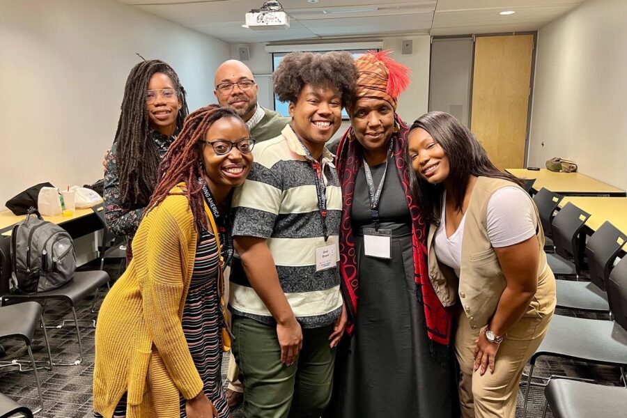 The group is gathered around Queen Cherice Harrison-Nelson who is on the right, with Toni Gordon leaning in next to her, Ural Grant on her right, Jessica Reed next to Ural, and in the back, Sharieka Botex and Marquis Taylor. They are in a conference room where the presentation on prioritizing joy in graduate education was given.