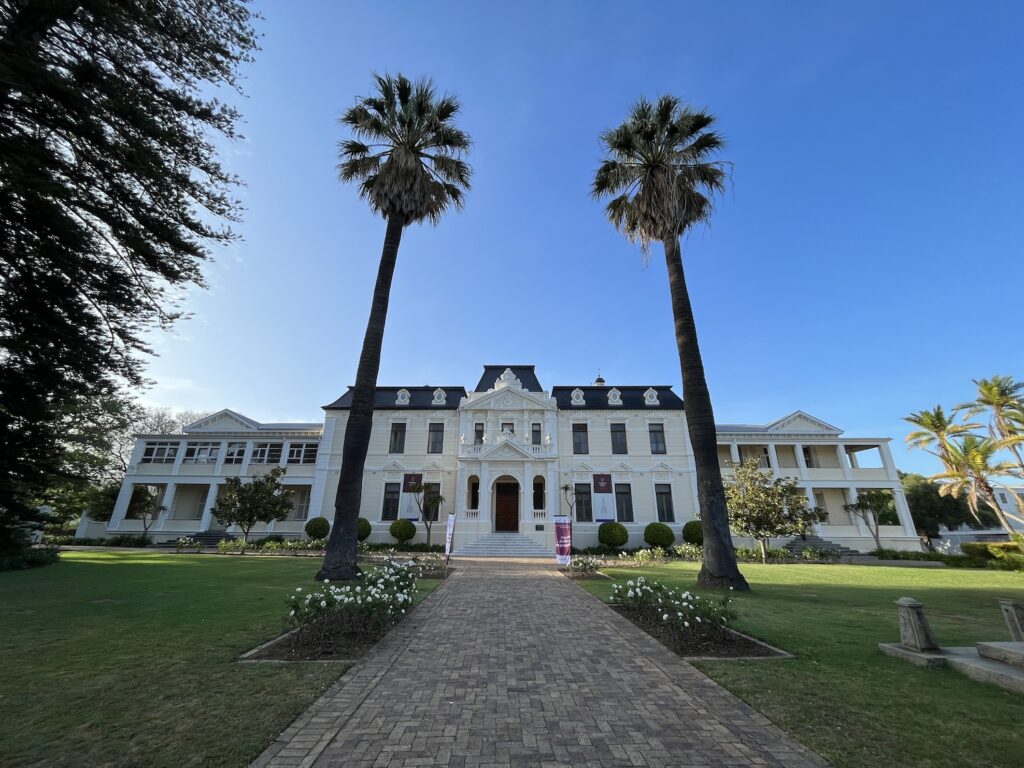 A grand, white, colonial-style building is at the end of a brick pathway framed by two huge palm trees. This is the entrance to the Faculty of Theology at Stellenbosch University. The picture is taken with a wide-angle lends and the trees tower overhead underneath a beautiful blue sky.