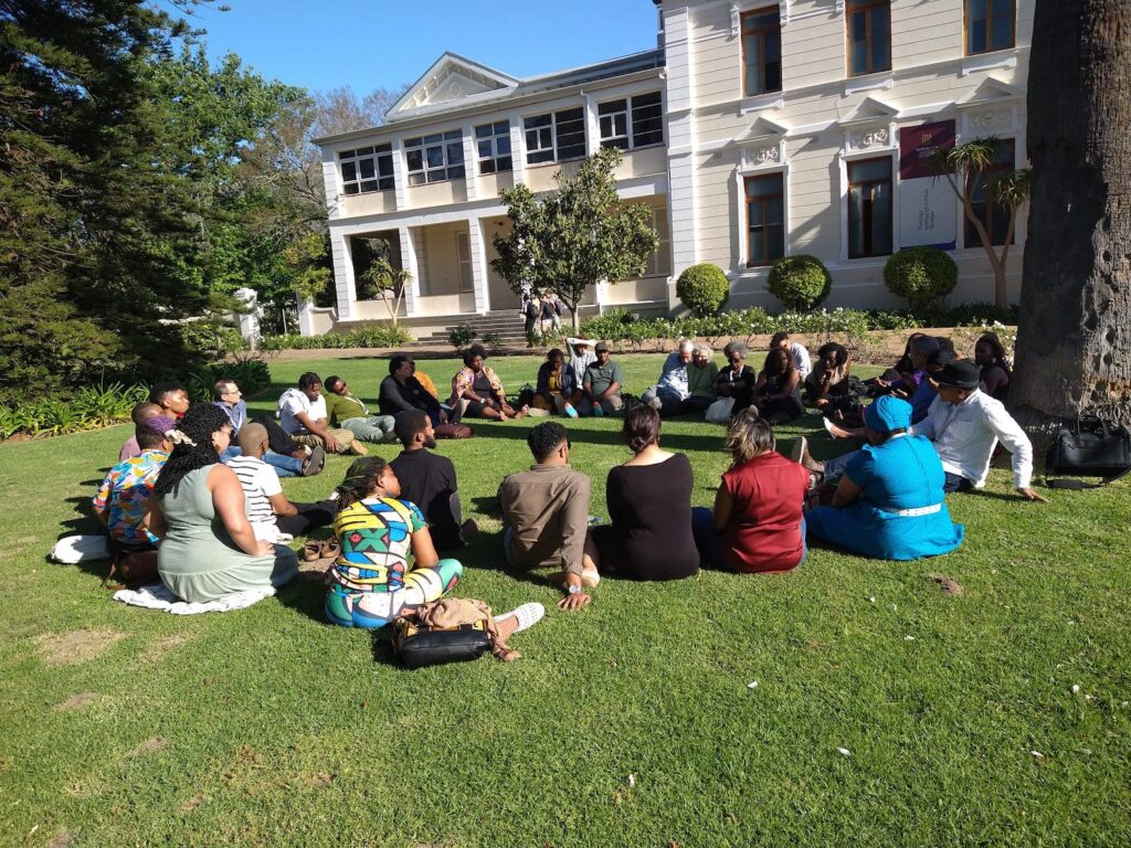 A group of more than 30 participants in the Ubuntu Dialogues conference sitting in a circle on green grass in front of the Faculty of Theology, a large, white, colonial-style building.