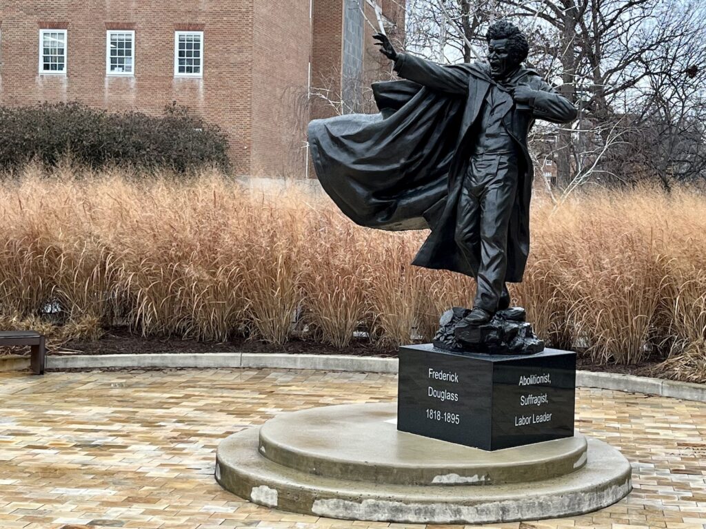 The Frederick Douglas statue on the University of Maryland campus depicts, as wikipedia puts it, "Douglass in the middle of a speech, with one arm outstretched, and a copy of his autobiography under the other arm." His mouth is open in a fiery speech. The statue is cast bronze on a black marble block with white text chiseled on front reading Frederick Douglas 1818-1895, on the side, white text is legible that reads: Abolitionist, Suffragist, Labor Leader. The figure is set off to the right of the image with a band of long, wheat colored grasses in the middle background, with three windows of the red brick colonial style building in the distant background.