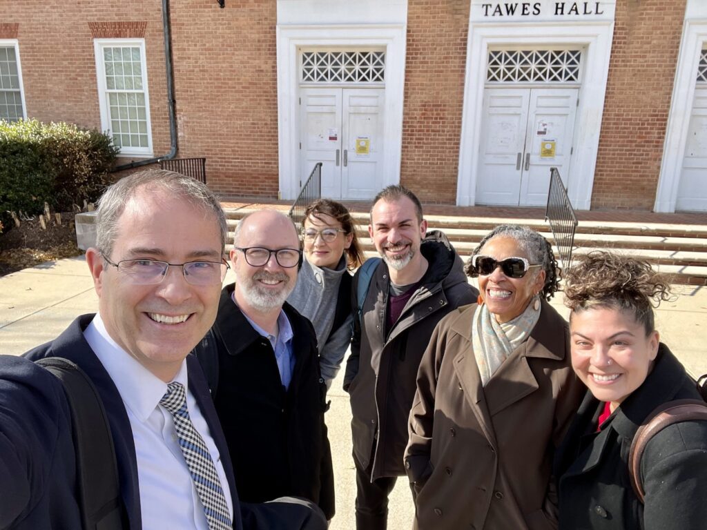 A selfie taking by Chris Long, who is on the left of the image with Jason Rhody, Nicky Agate, Simo Sacchi, Bonnie Thornon Dill, and Xhercis Méndez from left to right. The day is sunny, and Bonnie is wearing sunglasses. We are standing in front of the stairs and white doors leading into Tawes Hall, where we held the HuMetricsHSS workshop.