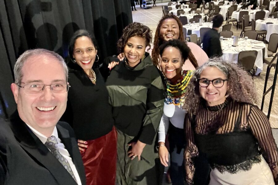 Selfie on stage at the Kellogg Center after the 43rd MLK Dinner at which LeConté Dill gave the keynote. The selfie is taken by Chris Long, who is on the left, with, from left to right, Ruth Nicole Brown, LeConté Dill, Chamara Kwakye, Gianina Strother, and Yvonne Morris. In the background are rows of empty round tables with white tablecloths and black chairs around them.