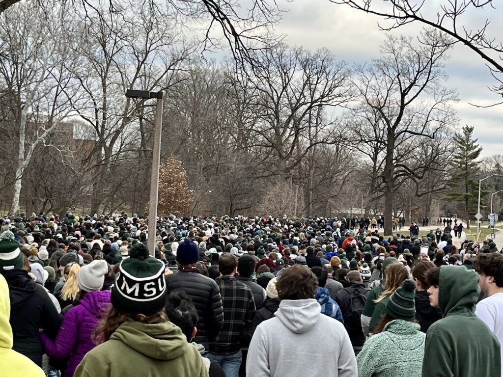 We are among a large crowd of people walking along the Red Cedar River trail toward the Rock on the MSU campus. The crowd extends off to the right along the river, as far into the distance. They are gathered for the Unity Walk to the vigil for the shootings on the MSU campus that occurred on Monday, February 13, 2023. People are in winter clothing and the trees are without leaves. A woman in a green and white MSU hat is in the foreground.