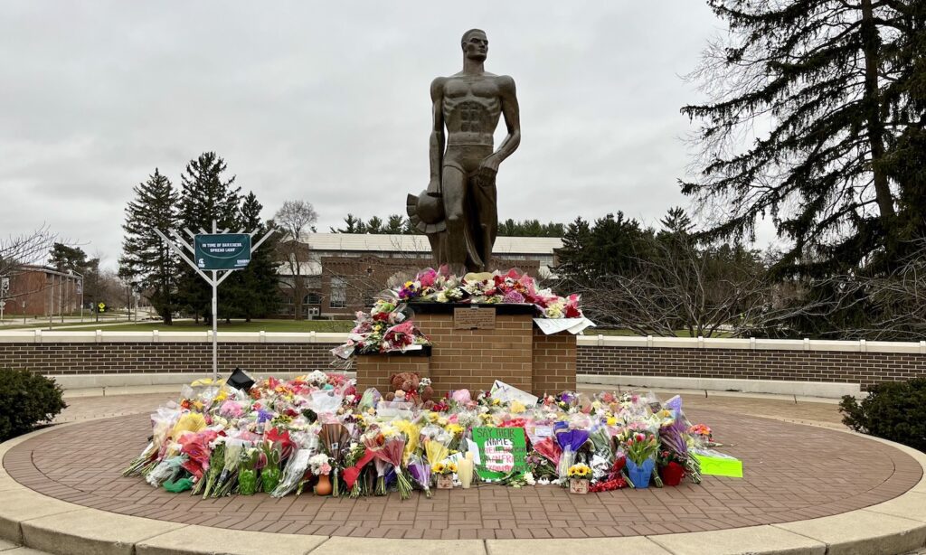 The Spartan Stature stands tall with flowers on its pedestal and on the platform at its base in honor of Alexandria Verner, Arielle Anderson, and Brian Fraser who died on February 13, 2023. There is a menorah in the background on the left.