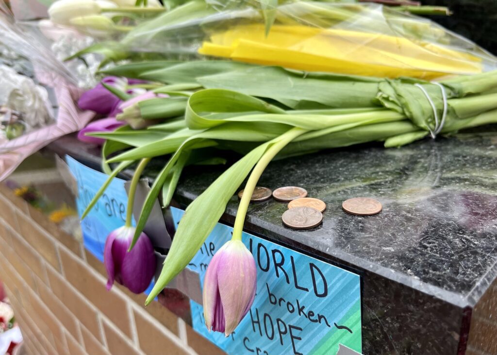 A corner of the black granite pedestal of the Spartan Statue has five pennies with purple tulips slumping over the edge to the left. There are blue and green card stock with the words World ... broken ... HOPE legible on it.
