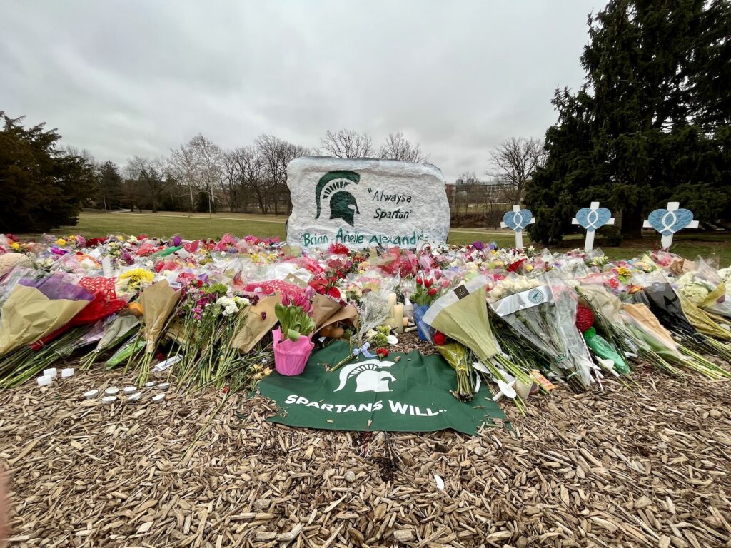 The Spartan Rock, painted white, with a green Spartan helmet, on the left and "Always a Spartan" painted on the front. The names of Brian Fraser, Arielle Anderson, and Alexandria Verner, are written in green letters along the bottom. In front of the rock are piles of flowers and a green and white Spartans Will flag laying on mulch front and center of the image. There are three crosses with hearts and fish symbols to the right of the rock.