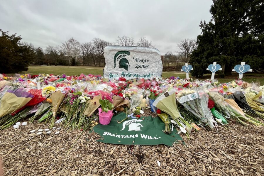 The Spartan Rock, painted white, with a green Spartan helmet, on the left and "Always a Spartan" painted on the front. The names of Brian Fraser, Arielle Anderson, and Alexandria Verner, are written in green letters along the bottom. In front of the rock are piles of flowers and a green and white Spartans Will flag laying on mulch front and center of the image. There are three crosses with hearts and fish symbols to the right of the rock.