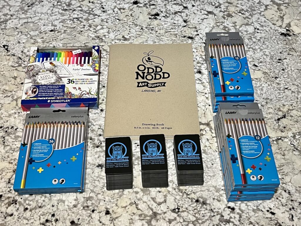 A collection of donations from Odd Nodd Art Supply and Blue Owl Coffee. The items are arranged in a rectangle, which Lamy colored pencil packs on the right side, a set of Odd Nodd Art Supply drawing books in the middle, which three stacks of Blue Owl Coffee cards - $625 worth in $5 increments, and on the left are Lost Ocean markers and more Lamy colored pencils.