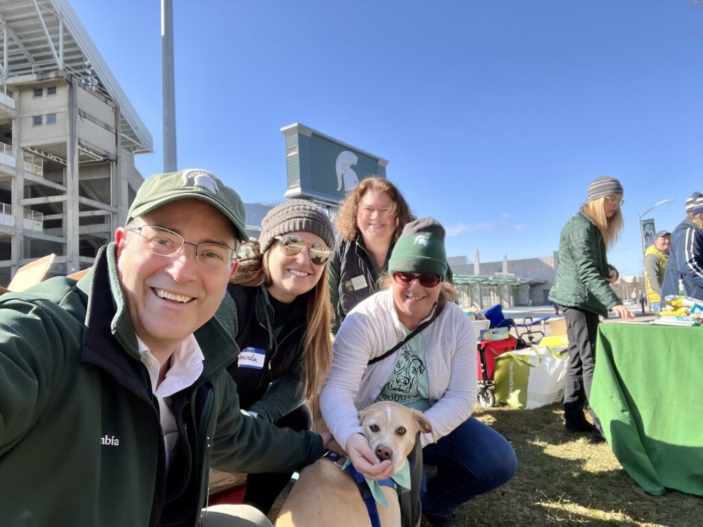 Selfie with Chris Long in Spartan green, Lupin the comfort dog is being given a treat to look at the camera, Amanda Ritter, Senta Goertler, and Bess German are in the selfie. Val Long is staffing the a table with treats for students in the background. In the background is Spartan Stadium with the Spartan logo large against a bright blue sky on Spartan Sunday to welcome students to campus after the February 13, 2023 shootings.