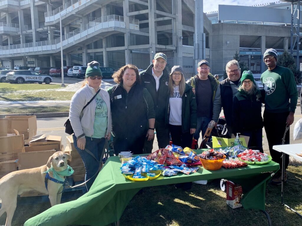 A posed picture of those who staffed the College of Arts & Letters and MSU Honors College table for Spartan Sunday with, from left to right, Lupin, the comfort dog, Senta Goertler, Bess German, Chris Long, Val Long, Dustin DeFelice, Tony Grubbs, Amy DeRogatis, and Andrew Wingard behind a table filled with candy and treats.