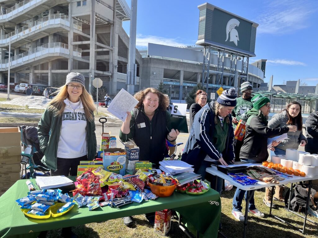 Val Long and Bess German staff the Spartan Sunday table filled with treats. Bess is holding up the MSU coloring book and Val is smiling. In the background is Spartan Stadium with the Spartan logo large against a bright blue sky on Spartan Sunday to welcome students to campus after the February 13, 2023 shootings.