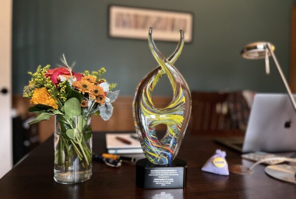 A dark wooden desktop has a bouquet of orange, white, and green flowers in a glass cylindrical vase on the left. In the middle is a multi-colored glass trophy in a double helix style on a black base on which the words are inscribed: "Michigan State University Institutional Champion Award for Community Engagement Scholarship Presented to Christopher P. Long, College of Arts and Letters and Honors College 2023." To the right, there is a small lavender bag of lavender with a yellow AAAS button on it. Blurred in the background on the right is an Apple MacBook Pro, the arm of a desk light and behind the trophy and flower are four fountain pens, a journal and a notepad.