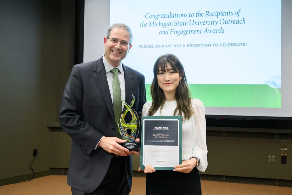 Chris Long and Krystal Jang pose next to each other with their Outreach and Engagement Awards. Behind them is a screen that reads: "Congratulations to the Recipients of the Michigan State University Outreach and Engagement Awards. Please join us for a reception to celebrate.