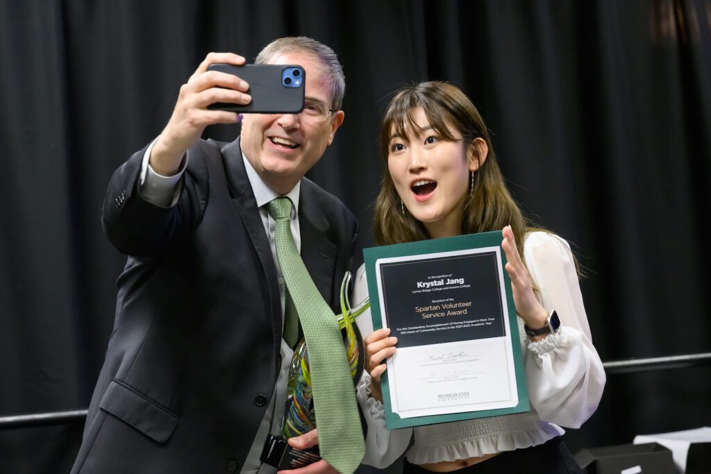 Chris Long holds an iPhone out to take a selfie with Krystal Jang, and Honors College student who is holding her Spartan Volunteer Service Award certificate. She is smiling for the camera, and you can only see one of Chris's eyes behind the phone. They are having fun.