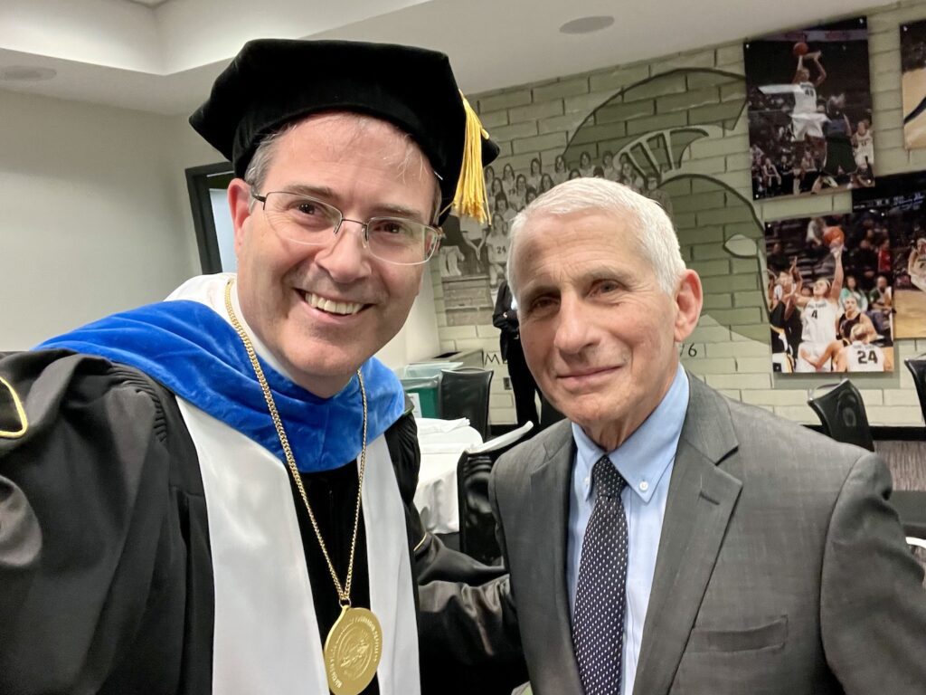 A selfie with Chris Long in academic regalia to the right of Dr. Anthony Fauci in the Breslin Center before the 2023 MSU Doctoral graduation ceremony. The wall behind them has a Spartan helmet and images of Spartan Basketball players in action.