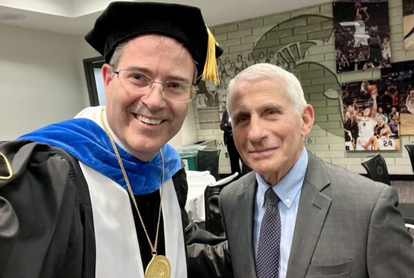 A selfie with Chris Long in academic regalia to the right of Dr. Anthony Fauci in the Breslin Center before the 2023 MSU Doctoral graduation ceremony. The wall behind them has a Spartan helmet and images of Spartan Basketball players in action.