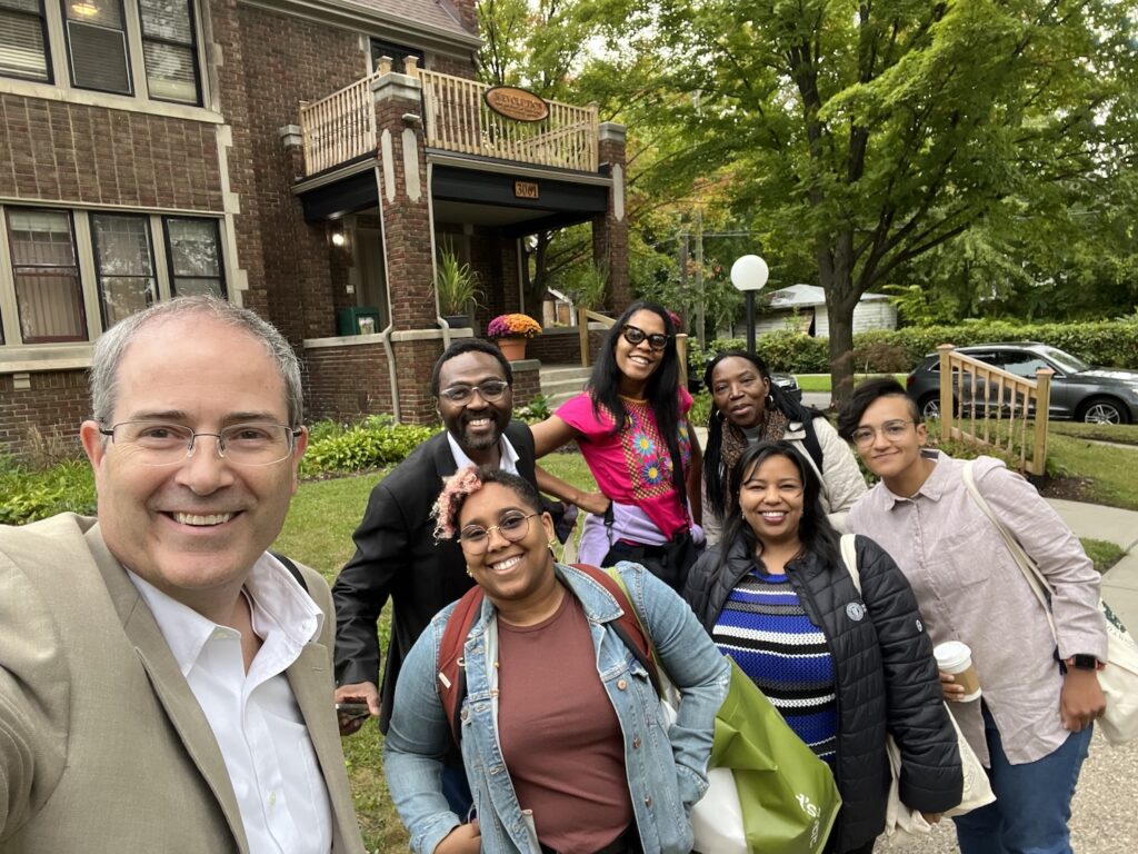 Selfie style image with Chris Long taking the picture with six colleagues from MSU and South Africa standing in front of the Boggs Center, a renovated brick house with a small front porch with a balcony as a roof.