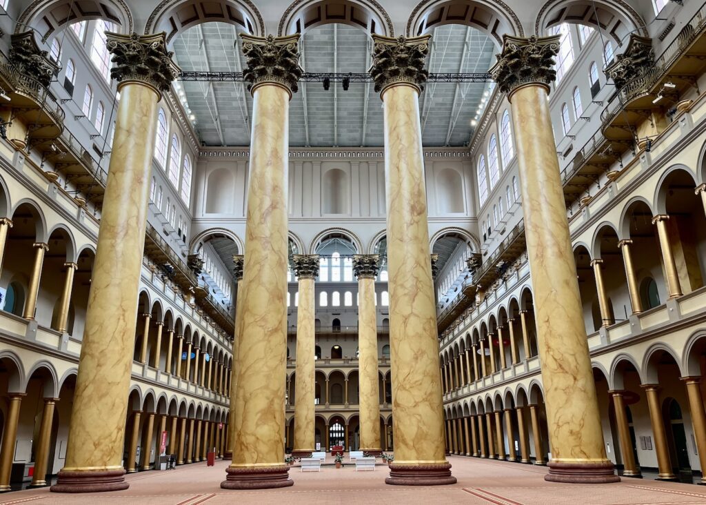 The atrium of the National Building Museum is an enormous space held up by four imposing Corinthian columns which stand before us. Two other columns are seen between the middle two columns further in the distance. The atrium has a rust colored rug. The columns are connected by arches between them. The atrium is surrounded by arched columns all around the first and second stories.