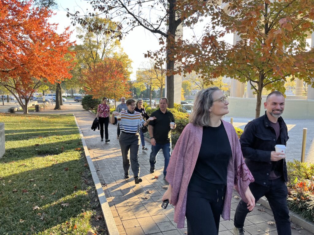 Members of the Coalition for the Future Academy walking (some with coffee) in Washington DC talking to each other along a path with trees in beautiful orange, green, and yellow folliage. They are two by two as they walk and talk.