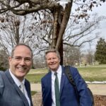Selfie with Chris Long and Kevin Guskiewicz in front of the Resilient Tree on MSU's campus. Long and Guskiewicz are in suit jackets with green ties and the Beaumont Tower is off in the distance on the left. The gnarled branches of the tree stretch out above them.
